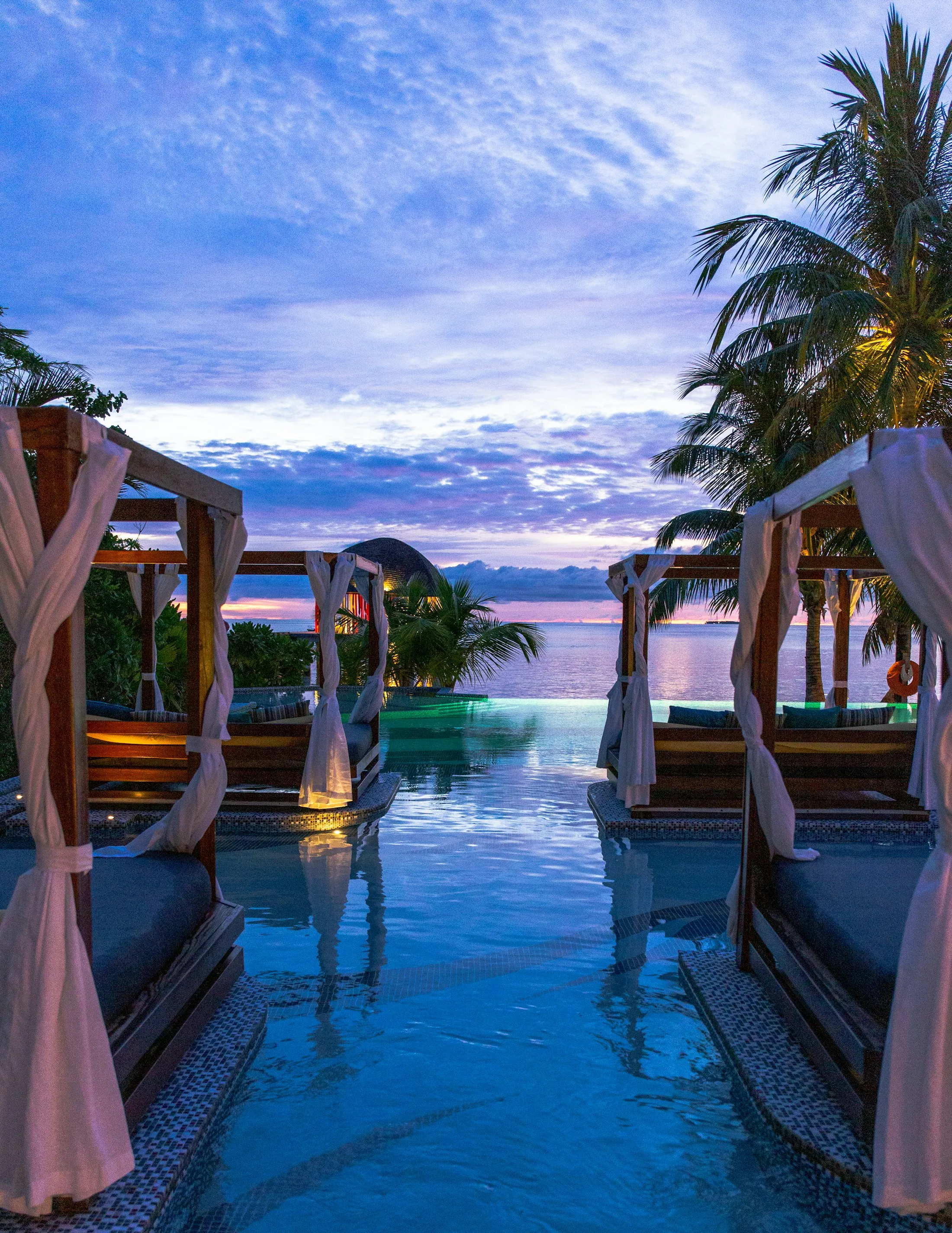 A Maldives resort pool with cabanas facing the sea at sunset