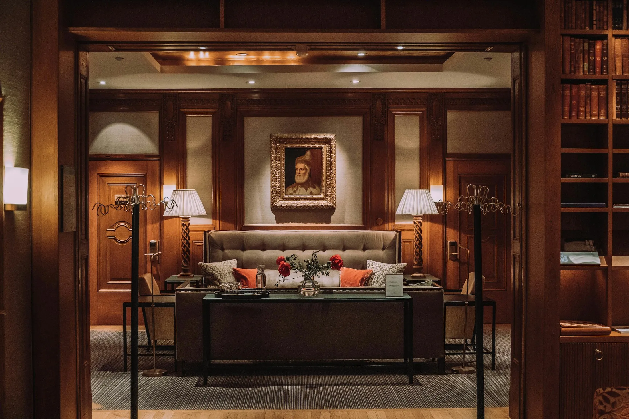 A wood-paneled luxury hotel sitting area with lamps, bookshelves, and a framed portrait