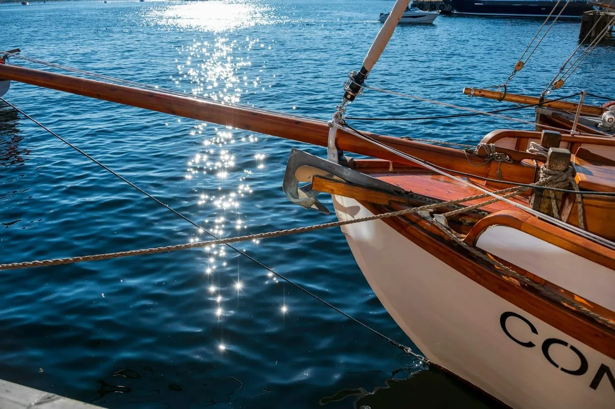 The varnished bow and bowsprit of a wooden sailboat moored in bright blue water